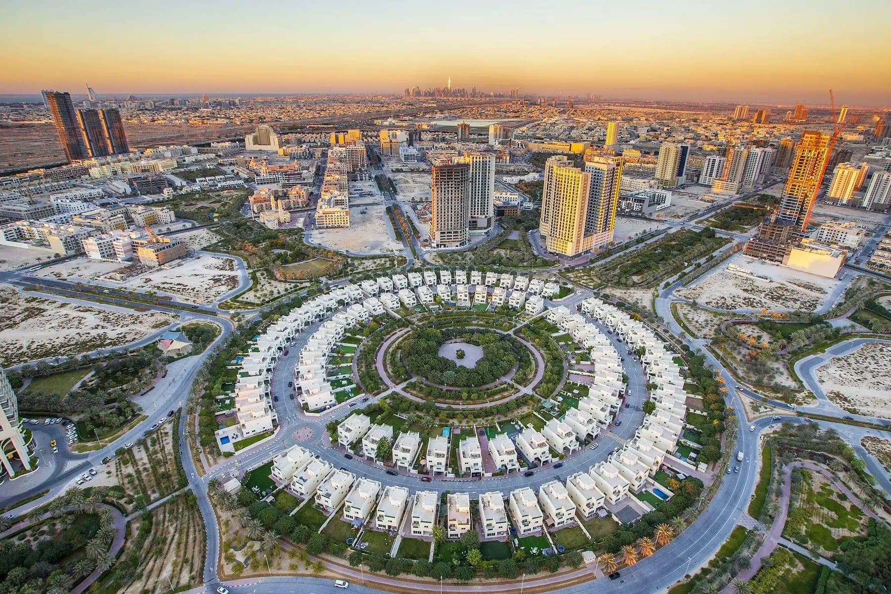 Aerial view of Jumeirah Village Circle at sunset, with its ring of villas around a central park and the Dubai skyline beyond